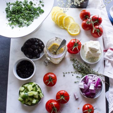 A jar of Greek Salad dressing surrounded by Greek Salad ingredients on a white marble and lemon slices