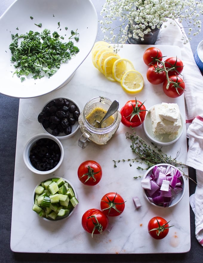 A jar of Greek Salad dressing surrounded by Greek Salad ingredients on a white marble and lemon slices