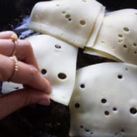 A hand placing the swiss cheese over the mushrooms over the cooked and ready burgers in a skillet