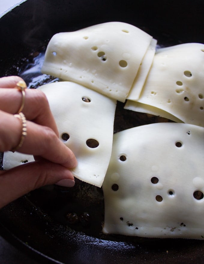 A hand placing the swiss cheese over the mushrooms over the cooked and ready burgers in a skillet