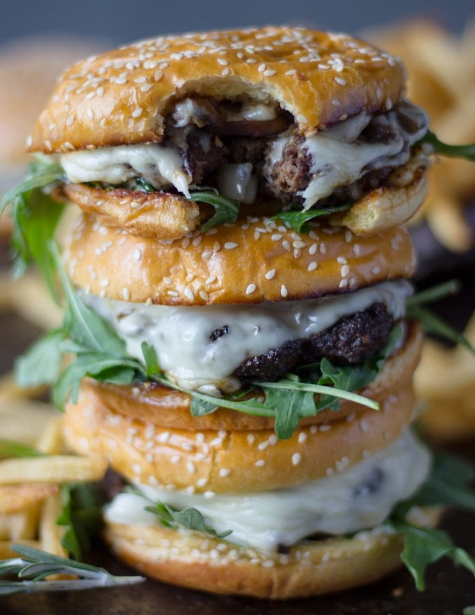 A stack of three mushroom swiss burgers with the top burger bitten up to show the melted swiss cheese, the mushrooms and the burger