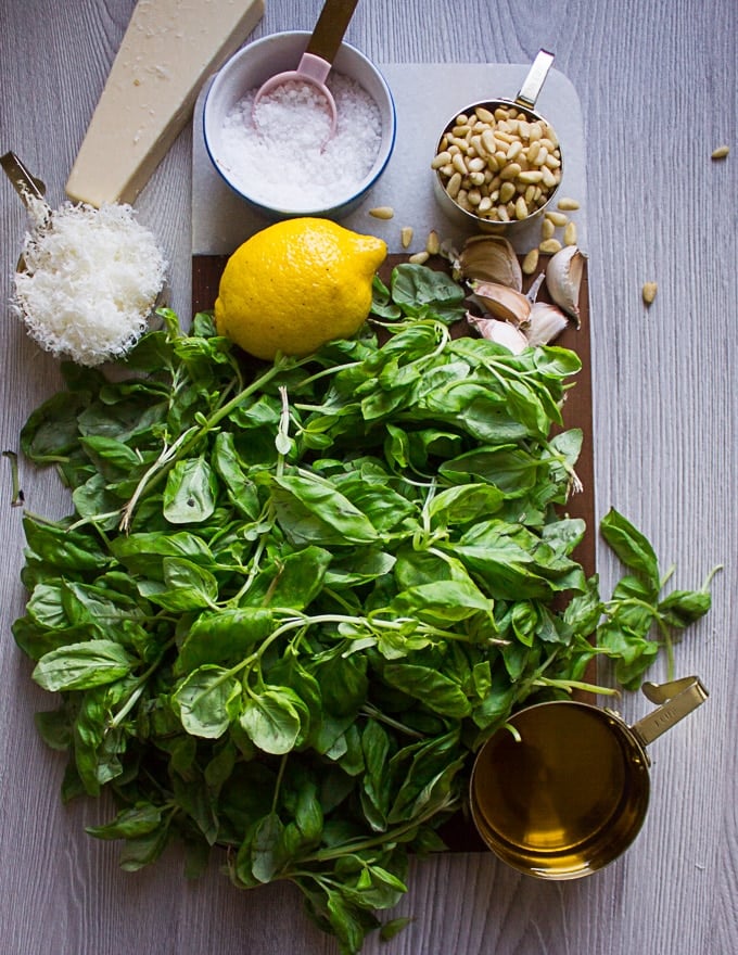 Ingredients for basil pesto over a wooden board such as fresh basil leaves, garlic, lemon, pine nuts, parmesan cheese salt and olive oil