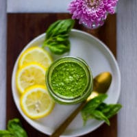 A jar of basil pesto over a whiite plate surrounded by lemon slices, fresh basil leaves and a spoon. All placed over a wooden board