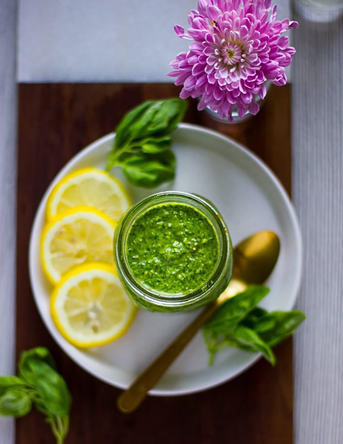 A jar of basil pesto over a whiite plate surrounded by lemon slices, fresh basil leaves and a spoon. All placed over a wooden board