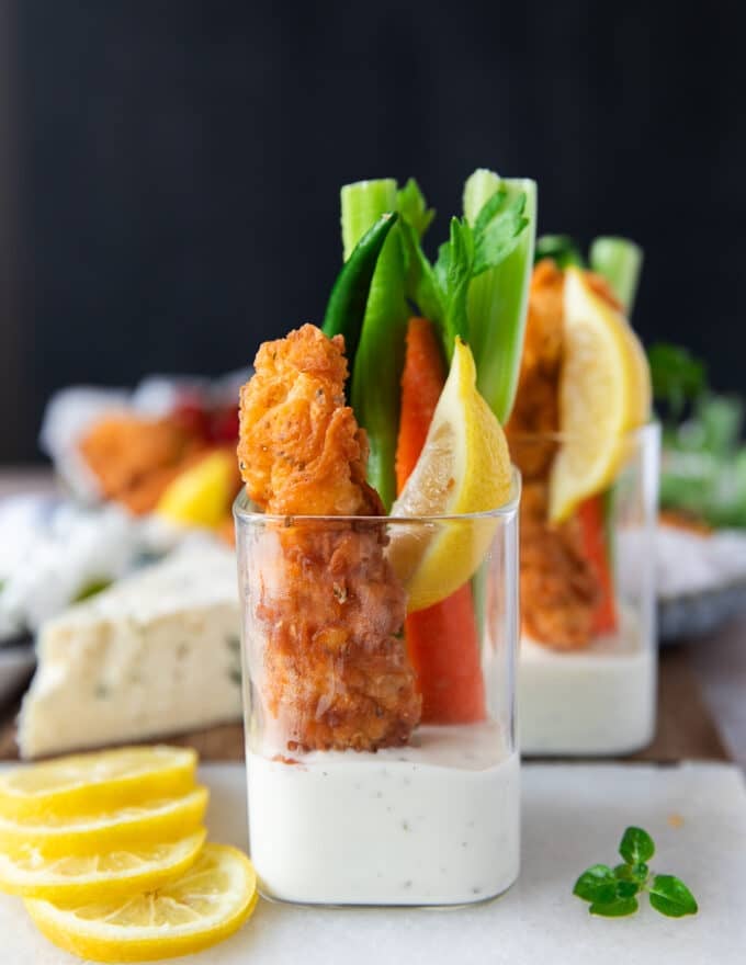 Close up of a cup with blue cheese dressing at the bottom, a chicken finger along with celery and carrots over the dressing in the cup