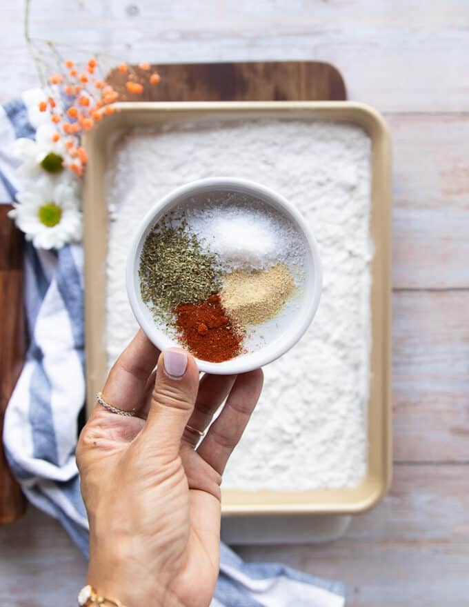 A hand holding a spice mix of paprika, garlic powder, oregano, salt and pepper and onion powder over a bowl of flour to make the dry coating