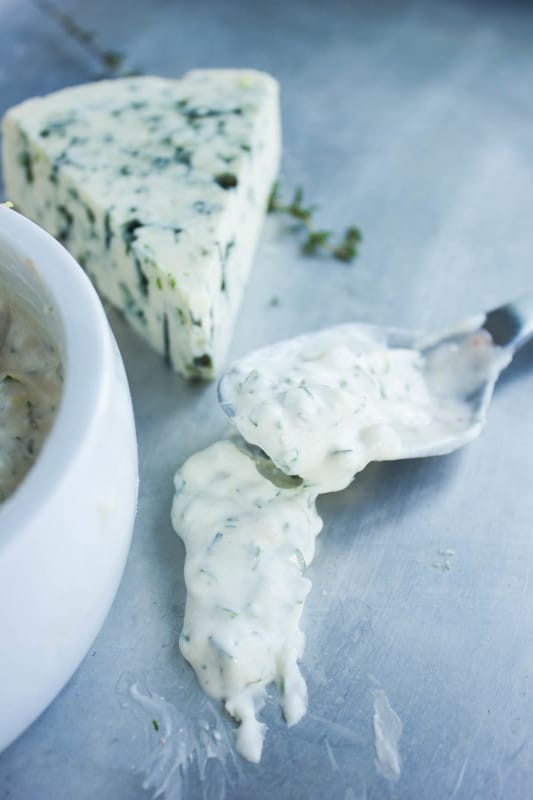 Blue cheese dip or bue cheese sauce spooned over a sheet by a spoon showing the consistency of it. A block of blue cheese in the background