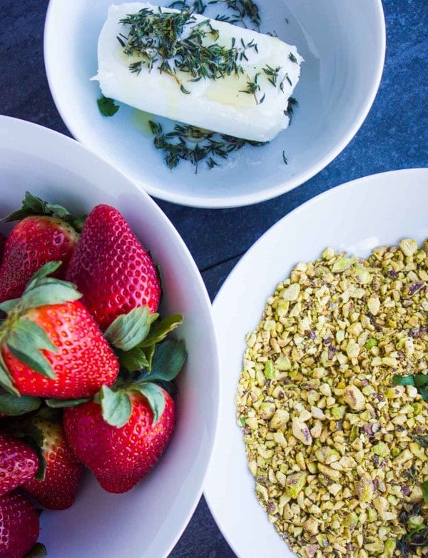 overhead of a plate of chopped pistachios next to a bowl of whole strawberries with a log of goat cheese in the background.