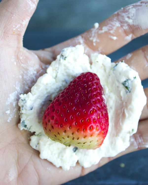 a strawberry sitting on a round circle of herbed goat cheese held in the palm of a hand, ready to be shaped into a ball