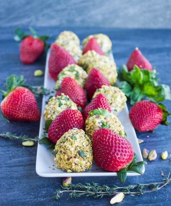Goat Cheese Pistachio Covered Strawberries and fresh strawberries lined up on a white rectangular plate with some fresh thyme twigs surrounding the plate.