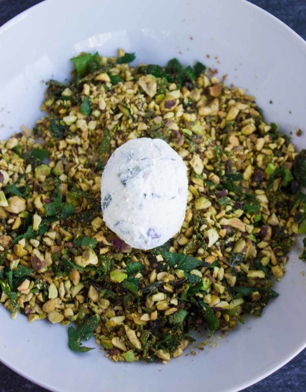 goat cheese ball sitting in a bowl of chopped pistachios and basil ready to be rolled