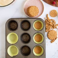 A muffin pan lined with baking sheets and a cookie pressed at the bottom of each paper to make the cheesecake base