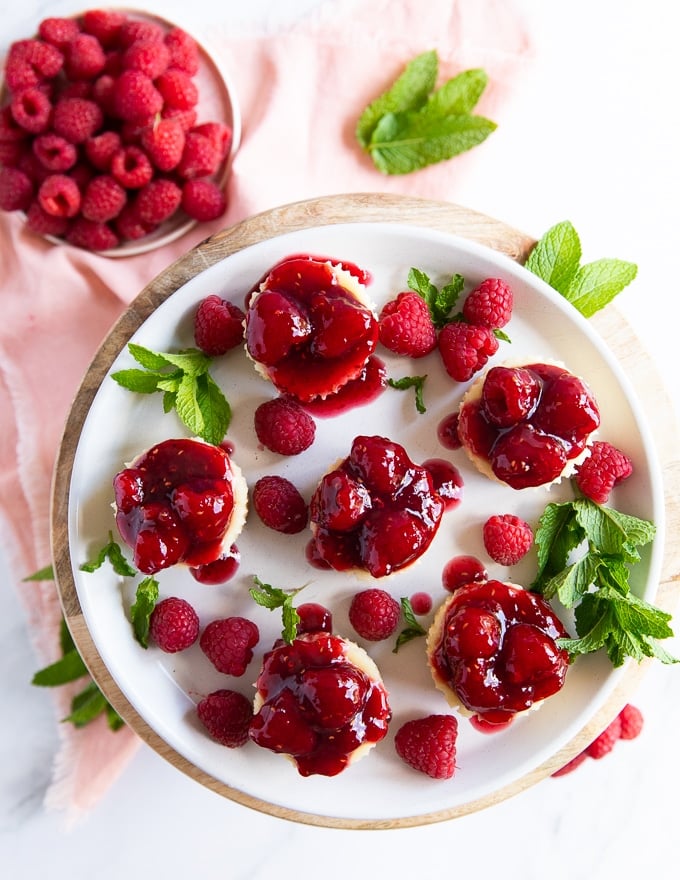 A full plate of mini cheesecakes surrounded by lots of fresh raspberries and fresh mint