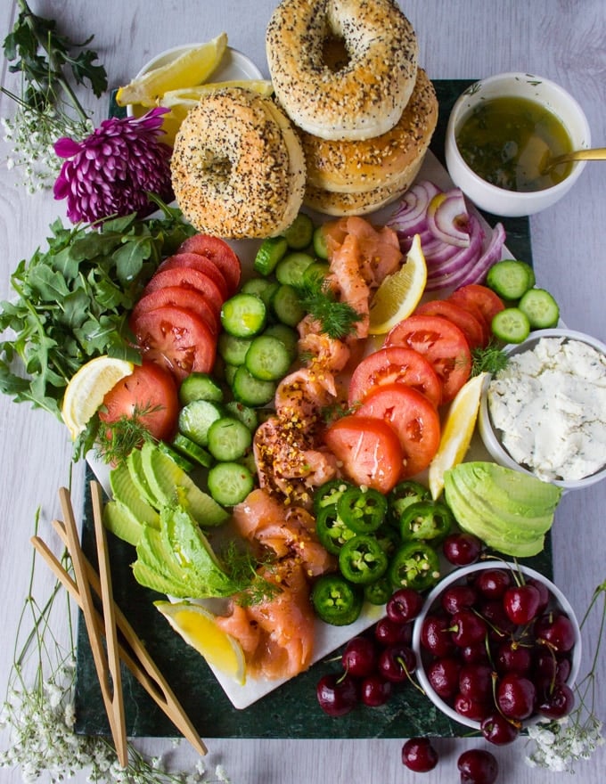 Chopped up and arranged smoked salmon platter on a marble board with sliced tomatoes, cucumbers, avocados, fruits, arugula, dill mustard sauce, herbed cream cheese, bagels.