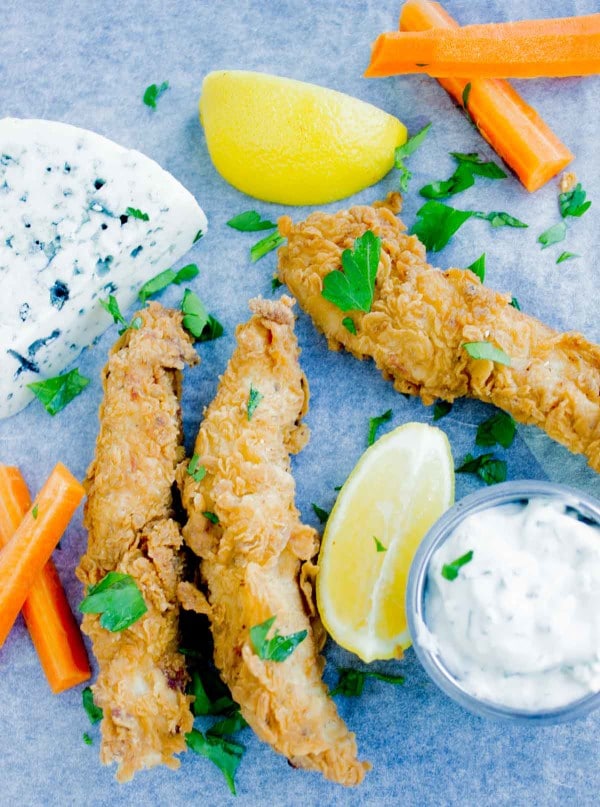 Spicy Fried Chicken Fingers arranged on a blue rustic tabletop with a dollop of blue cheese sauce and some carrot sticks on the side.