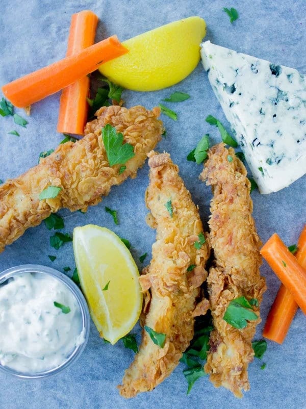 Spicy Fried Chicken Fingers arranged on a blue rustic tabletop with a dollop of blue cheese sauce and some carrot sticks on the side.
