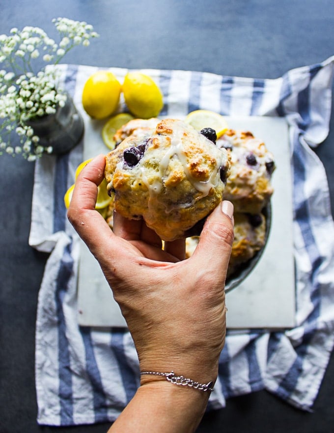 A hand holding a glazed drop biscuits with blueberries and lemon over a plate of drop biscuits