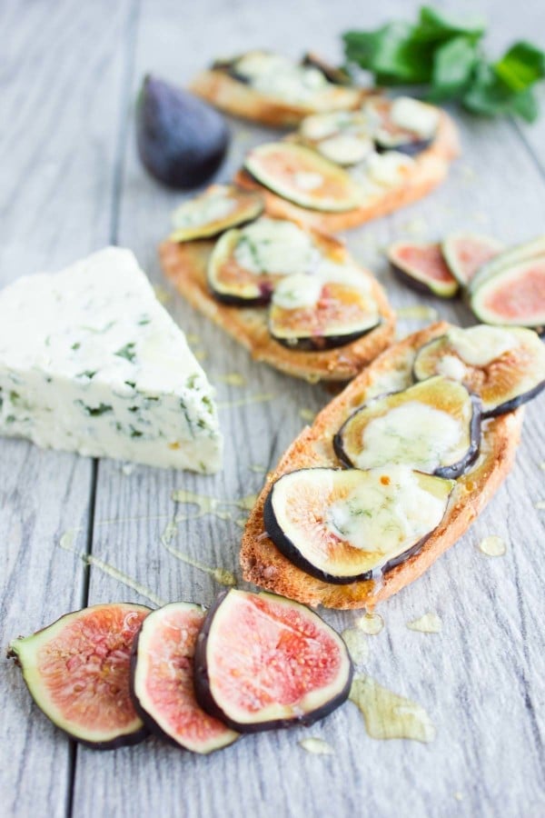 Fig Blue Cheese Honey Crostini drizzled with honey, served on a rustic table with a wedge of blue cheese and some figs in the background