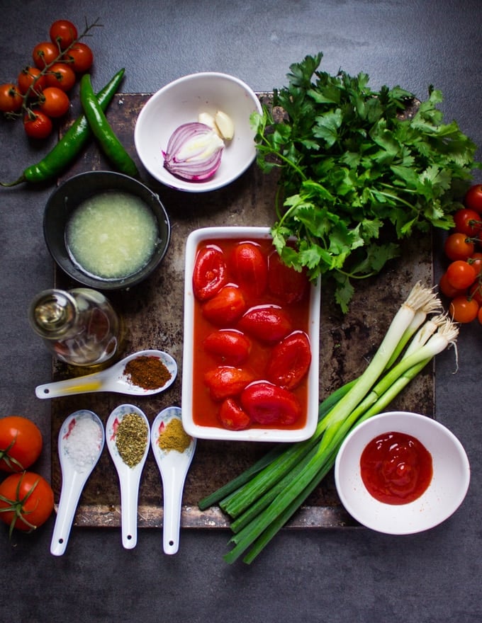 ingredients of homemade salsa on a board. a bowl of tomatoes, scallions, lime juice in a bowl, seasoning in small spoons, scallions, fresh parsley and cilantro, onions, garlic, ketchup and jalapeno