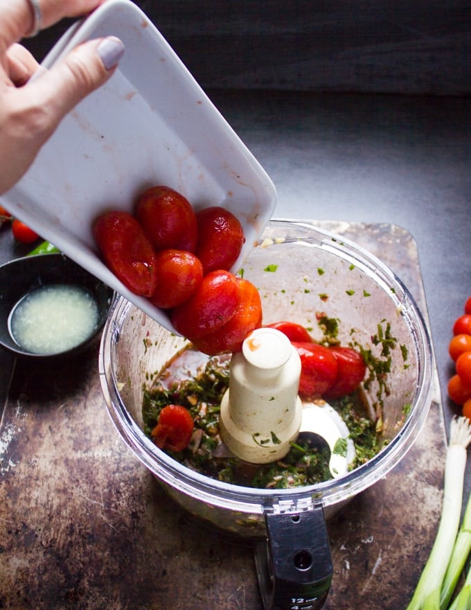 a hand pouring the tomatoes in a bowl of food processor where other ingredients have already been processed.