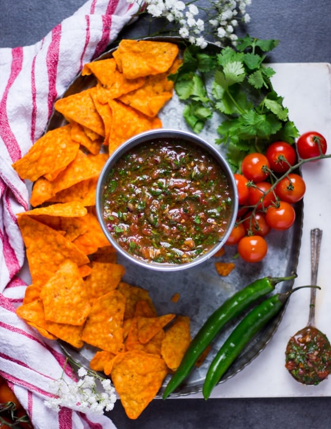 a metal bowl of salsa surrounded by chips, fresh tomatoes, jalapeno and cilantro over a tray