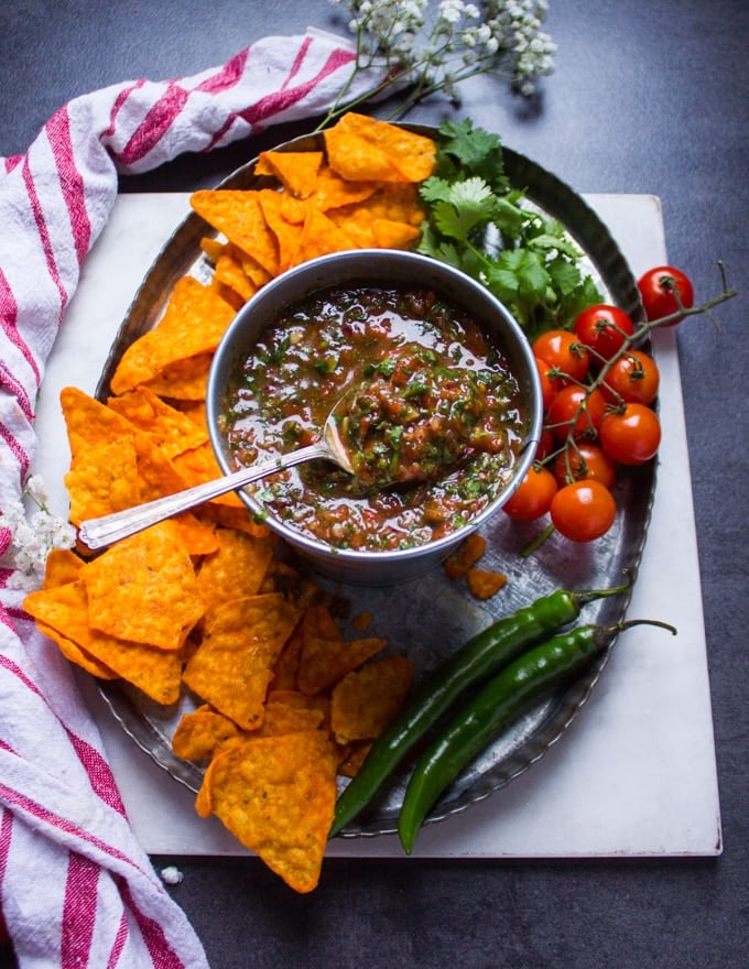 A spoon over a bowl of homemade salsa scooping some out. Surrounded by chips, jalpeno, a tea towel, cilantro and fresh tomatoes