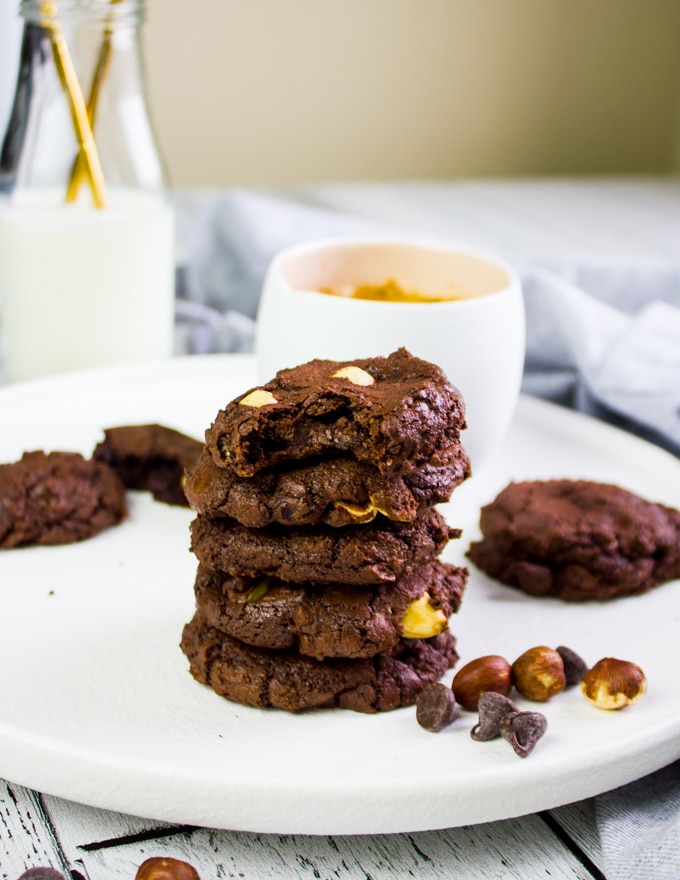 A plate with piled up brownie cookies and a cup of coffee, some hazelnut and chocolate chips scattered around it