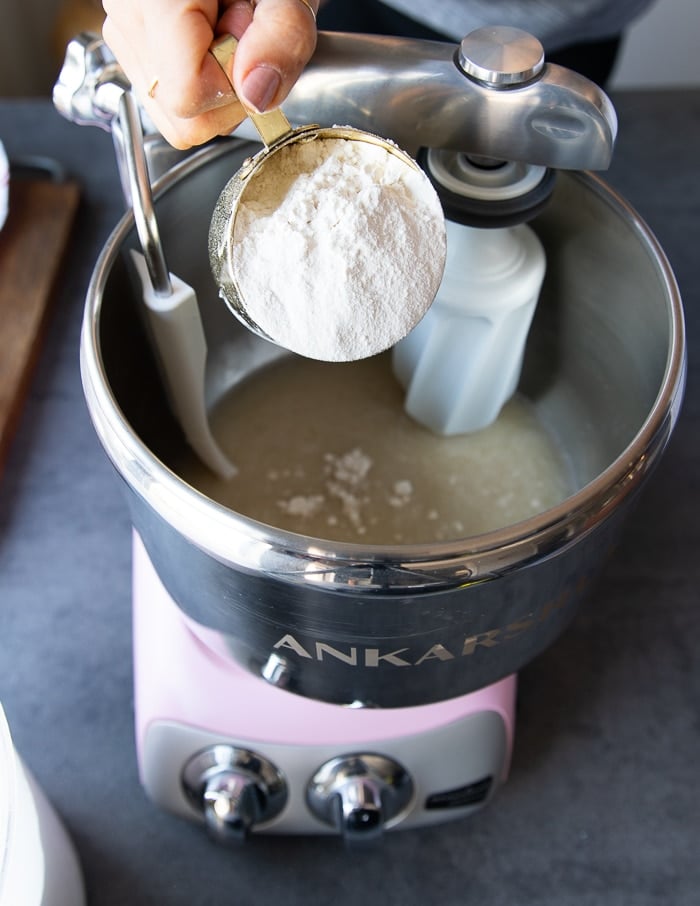 A hand pouring in the flour gar4dually into a stand mixer to make the pizza dough recipe