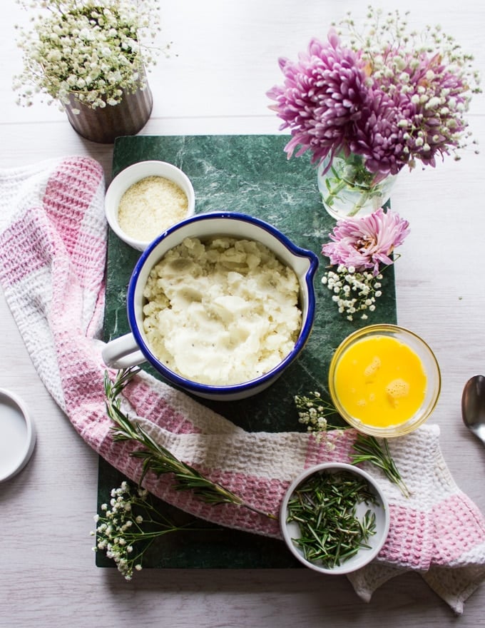 Ingredients for the potato pancakes, a bowl of leftover mashed potatoes, an egg in a bowl, fresh rosemary in a bowl, grated parmesan cheese in a bowl surrounded by fresh flowers