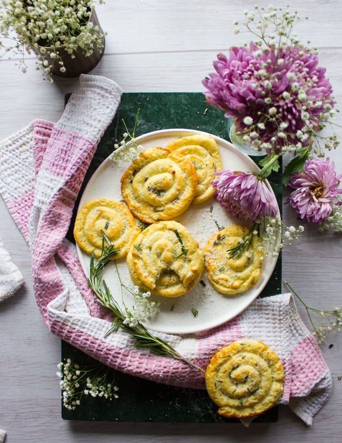 Top view of a plate with potato pancakes sprinkled with fresh rosemary and extra parmesan cheese, surrounded by a tea towel and fresh flowers