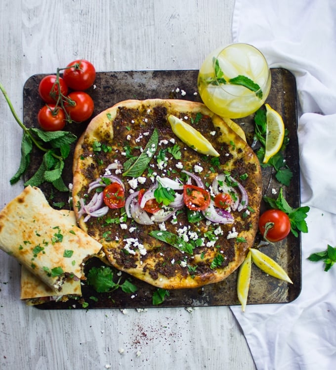 Several Turkish Pizzas served on a large sheet, some of them flat while others rolled up, on the side some tomatoes and juice, lemon slices and herbs