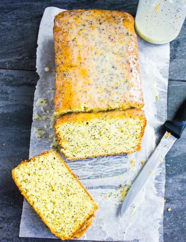 Overhead shot of a sliced Citrus Poppy Seed Loaf cake with Yogurt Glaze on parchment paper with a jar of glaze in the background.