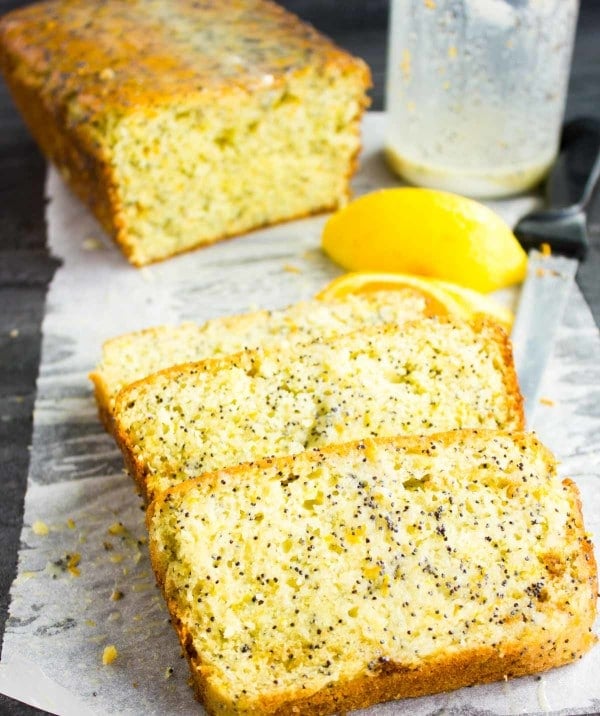 Close-up of three slices of Citrus Poppy Seed Loaf with Yogurt Glaze on white parchment paper with lemons and half a loaf cake in the background.