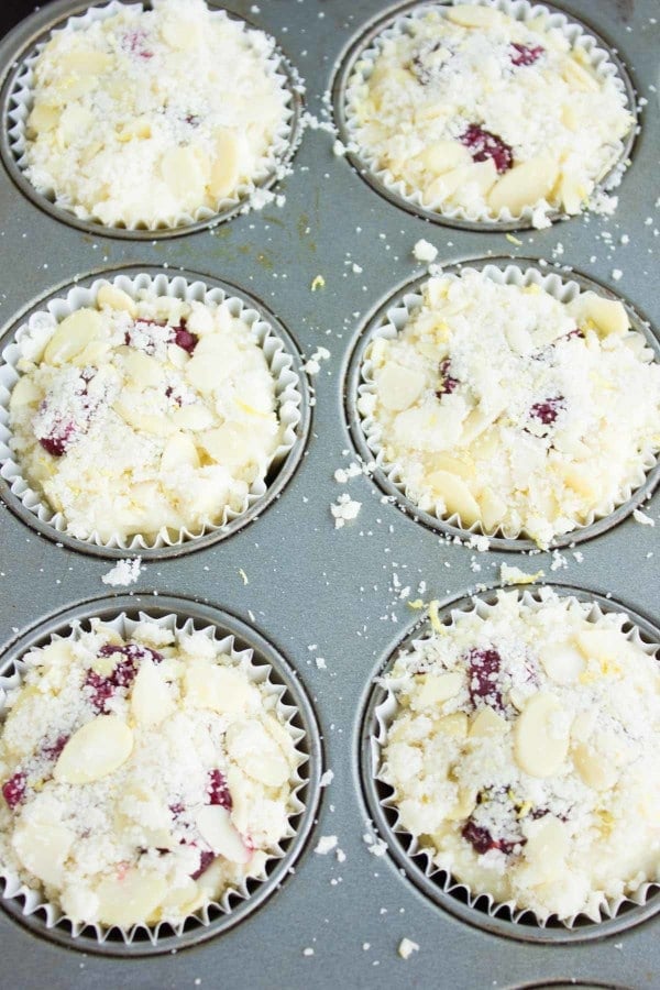 a paper-mold lined muffin tray filled with unbaked muffin batter topped with fresh raspberries and almond crunch topping.