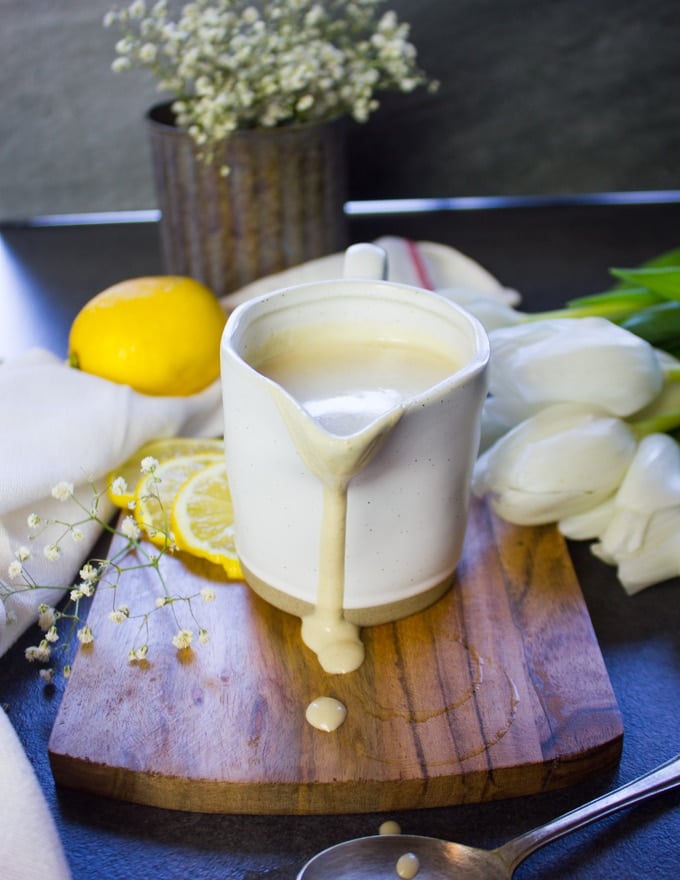 The tahini sauce in the pouring jar dripping over a wooden board surrounded by lemon slices. 