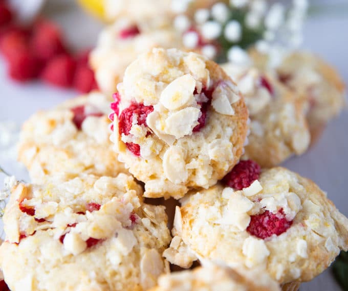 stacked up raspberry muffins over one another with fresh rasberries around them and some lemons in the background on a white board