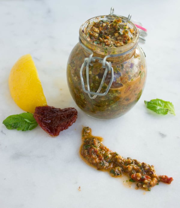 Sun Dried Tomato Pesto in a glass jar with some lemon, sun-dried tomatoes and basil leaves on the side