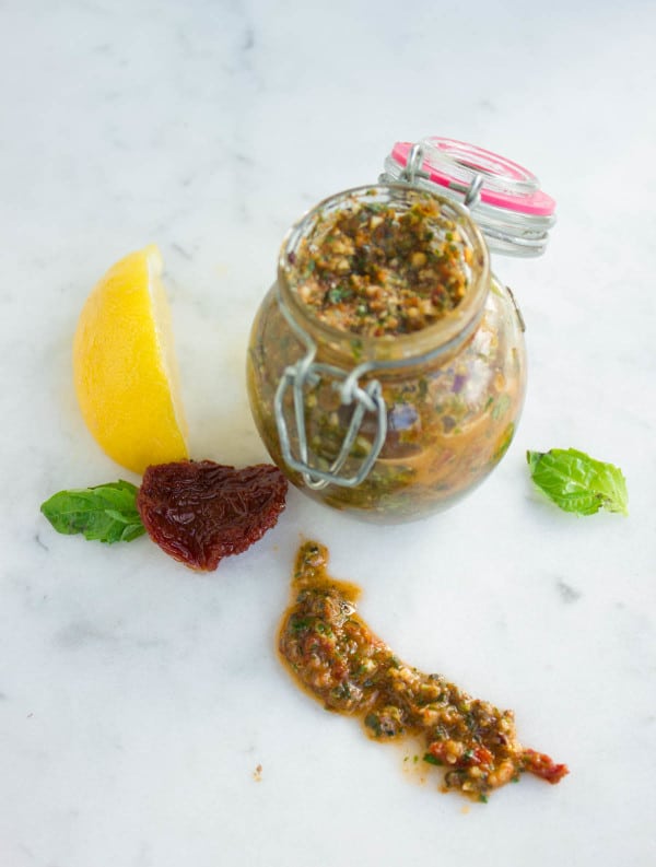 overhead shot of Sun-Dried Tomato Pesto in a glass jar on a white tabletop with some pesto ingredients on the side