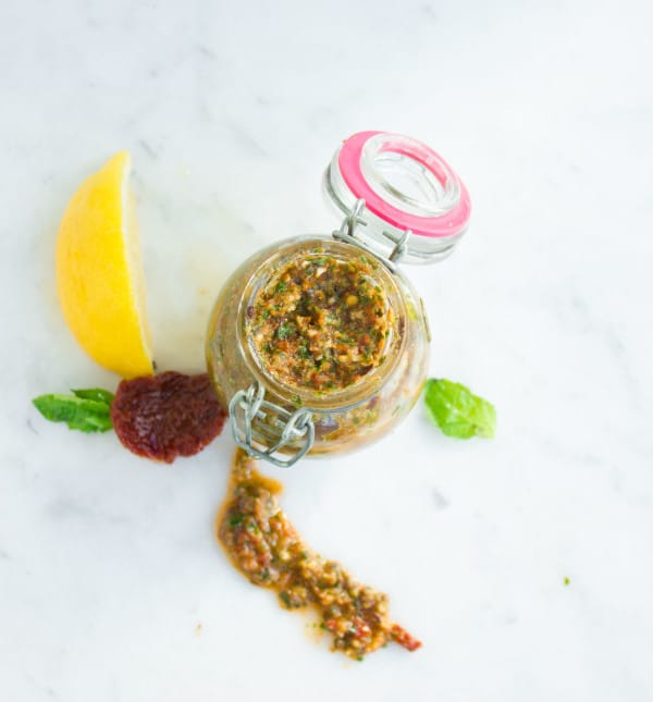 overhead shot of Sun-Dried Tomato Pesto in a glass jar on a white tabletop with some pesto ingredients on the side