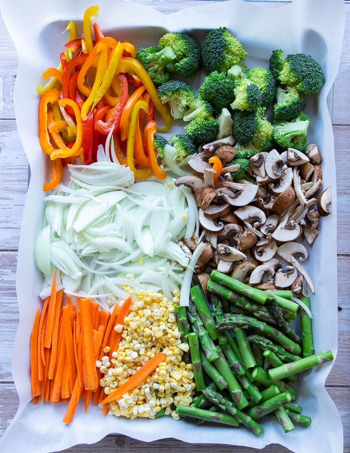Spring veggies on a sheet pan lined with parchment: yellow peppers, broccoli, onions, mushrooms, carrots, corn, and asparagus.