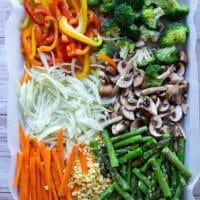 Seasoned and ready Spring veggies on a sheet pan lined with parchment: yellow peppers, broccoli, onions, mushrooms, carrots, corn, and asparagus.