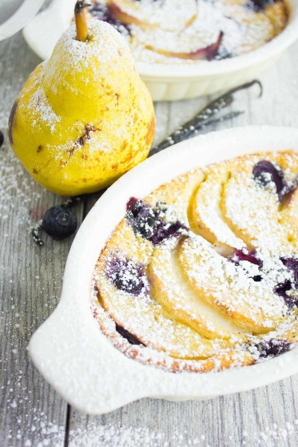individual Pear Blueberry Vanilla Clafoutis dusted with icing sugar with a pear and vanilla bean on the side