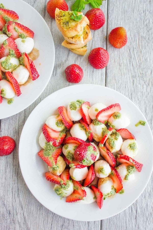 Overhead shot of Strawberry Caprese Salad drizzled with pesto, served on a white plate with baguette slices and whole strawberries in the background.