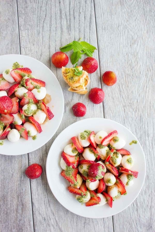 Overhead shot of Strawberry Caprese Salad drizzled with pesto, served on two white plates with baguette slices and whole strawberries in the background.