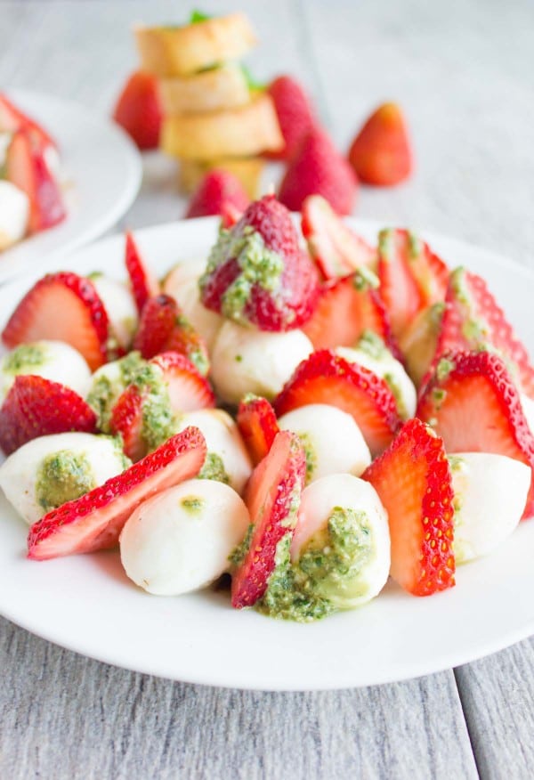 Close-up of Strawberry Caprese Salad drizzled with pesto, served on a white plate with baguette slices and whole strawberries in the background.