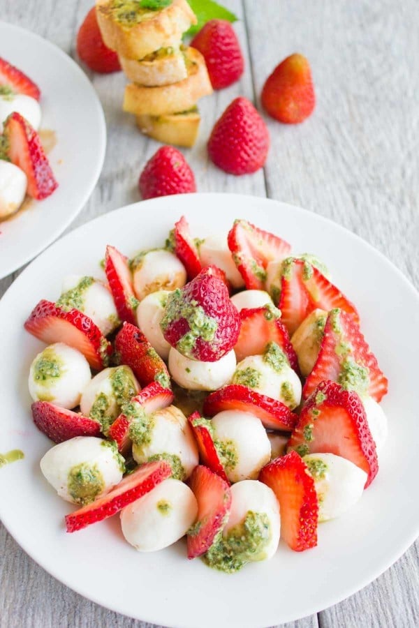 Overhead shot of Strawberry Caprese Salad drizzled with pesto, served on a white plate with baguette slices and whole strawberries in the background.