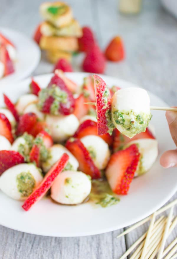 a mini mozzarella ball and a slice of strawberry smothered with green pesto skewered on a toothpick with a plate of Strawberry Caprese Salad in the background.