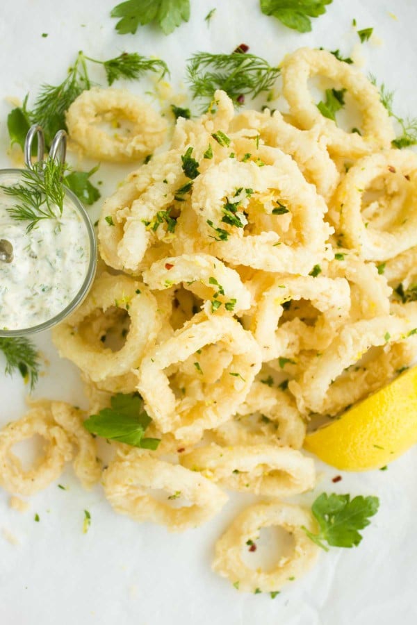 Zesty Fried Calamari rings arranged on a white tabletop with a side of tartar sauce and some fresh dill