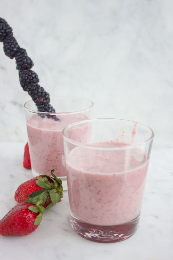 2 glasses of Banana Berry Avocado Chia Smoothie on a white kitchen counter with a blackberry skewer and some fresh strawberries in the background