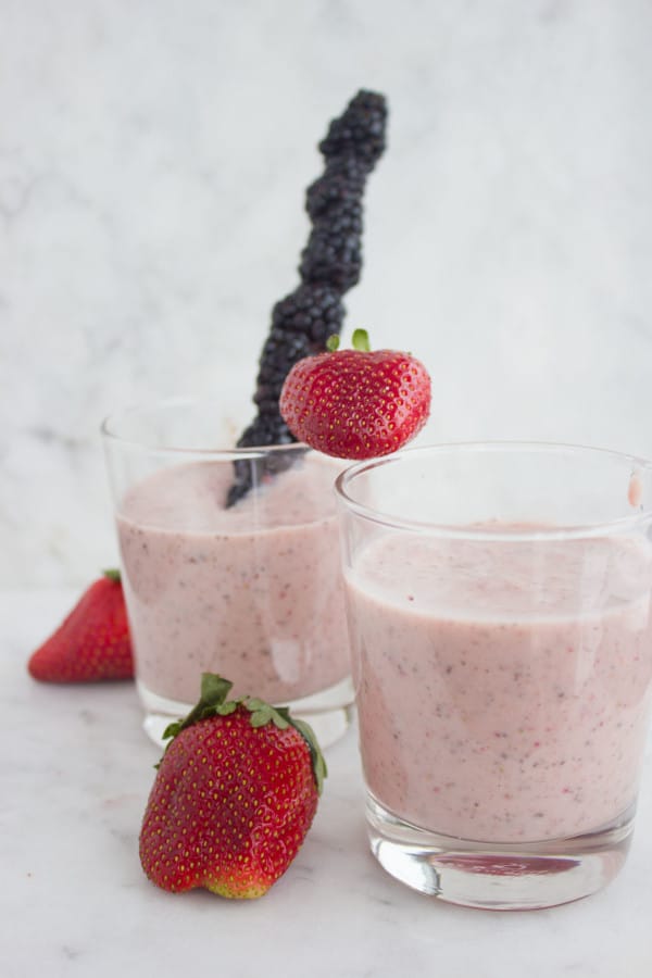 2 glasses of Banana Berry Avocado Chia Smoothie on a white kitchen counter with a blackberry skewer and some fresh strawberries in the background